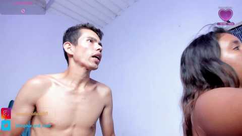 Video of a shirtless, fit young man with short dark hair, standing beside a woman with long dark hair, both looking surprised. Minimalist white room background.