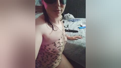 A video shows a fair-skinned woman with dark hair, wearing a leopard-print bodysuit, sitting on a bed in a dimly lit room. She has black sunglasses and a black strap around her neck.