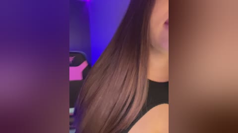 Video of a young woman with long, straight brown hair, wearing a black tank top, partially cropped. Background features a purple light and a gaming chair.