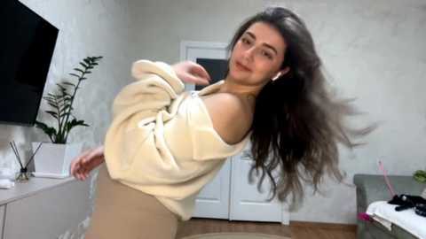 Video of a young woman with long brown hair, wearing a cream off-shoulder sweater and beige pants, standing in a modern, minimalist room with white walls, a TV, and potted plants.