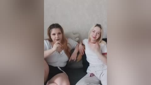 A video of two young women with fair skin and long hair, wearing casual clothes, seated on a couch in a dimly lit room.