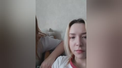 A video of a young Caucasian woman with long blonde hair, lying on a bed, partially visible behind a partition. She has a neutral expression.