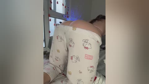 A video of a woman in white Hello Kitty pajama pants, bent over a bed, showing her buttocks. The background features a window with fairy lights and a white wall.