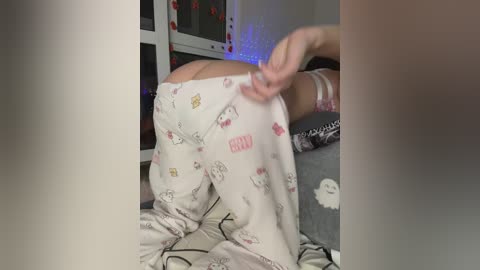 Video of a person in pajamas with a cartoon print, pulling up pajama bottoms, showing buttocks, in a bedroom with a window and blue fairy lights.