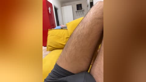 Video of a man's hairy legs in gray shorts, reclining on a yellow sofa, with a red refrigerator and white wall background.