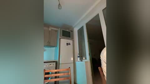 Video of a small, dimly lit kitchen with white appliances, wooden chair, and a partially open door leading to a dark hallway.