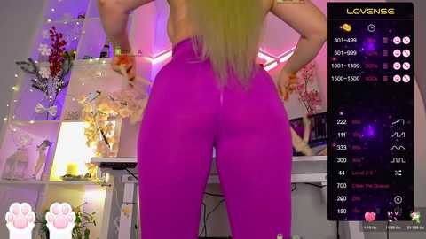 Video of a woman with long blonde hair wearing tight, bright pink leggings, standing with hands on hips, in a cozy, softly lit room with floral decor and a video recording screen.