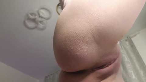 Video of a close-up, slightly tilted view of a light-skinned woman's left breast and nipple, with a tattoo on her lower abdomen. Background features a white ceiling with a decorative light fixture and part of a grey curtain.