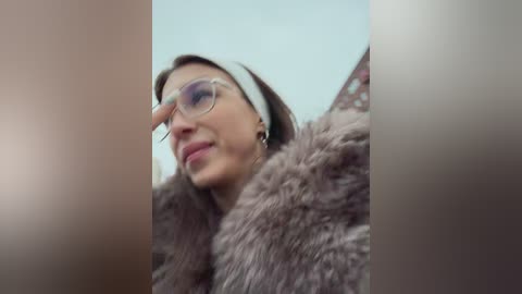 Video of a woman with short brown hair, wearing clear glasses, a beige headband, and a fur coat, viewed from a low angle, with blurred background.