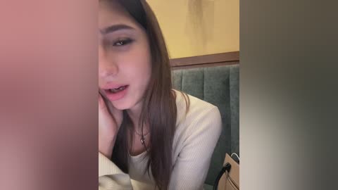 A video captures a young woman with long brown hair and fair skin, wearing a beige sweater, resting her chin on her hand, looking pensive. She sits in a teal upholstered chair in a dimly lit room with yellow walls.