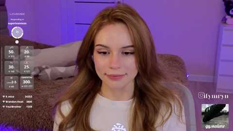 Video of a young, light-skinned woman with long, wavy brown hair, wearing a white shirt, sitting on a bed with a beige blanket. Background shows a purple-lit room, social media icons, and a close-up of a man's face in the bottom right corner.