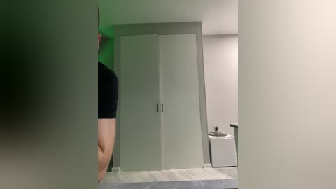 Video of a person with fair skin and a black T-shirt, standing in front of a mirrored closet door in a minimalist bathroom with a white toilet and a green accent wall.