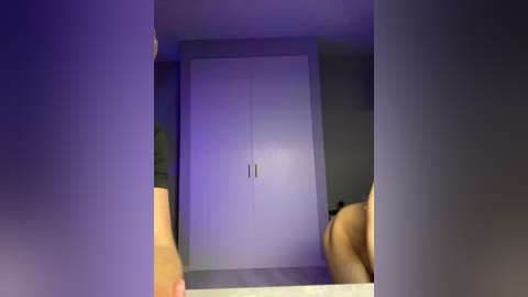 Video of a modern, minimalist bedroom with a white wardrobe in the background, illuminated by purple light. The image is framed by the viewer's outstretched arms, suggesting a selfie perspective.
