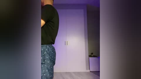 A video of a man wearing dark jeans and a green shirt, standing in a dimly lit room with a white wardrobe and a small nightstand.