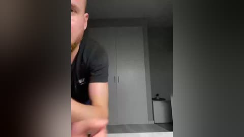 Video of a bearded man in a black t-shirt, partially visible, standing in a minimalist, modern bathroom with white walls, a white toilet, and a mirror.