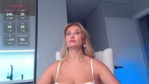 A video of a blonde woman with long hair, wearing a white dress, sitting in a modern room with a temperature display showing 29\u00b0C.