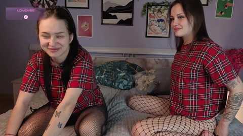 Video of two young women with pale skin, straight brown hair, and tattoos, wearing red plaid shirts and fishnet tights, sitting on a bed with blue pillows and green walls adorned with art.