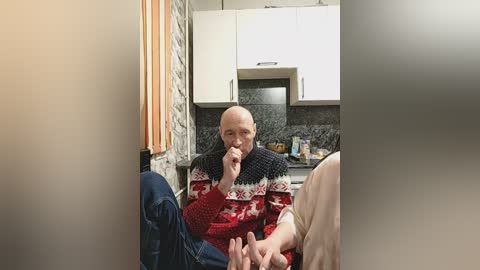 Video of a bald, middle-aged man in a red sweater with white and black patterns, eating a sandwich in a small, modern kitchen with white cabinets and dark countertops.