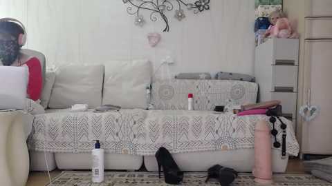 Video of a messy living room with a white couch, white wall art, and scattered items on the floor, including shoes, a can, and a doll.