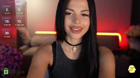 Video of a smiling woman with long black hair, wearing a black tank top and a cross necklace, in a dimly-lit, modern room with a purple flower arrangement and a TV screen displaying time and date.