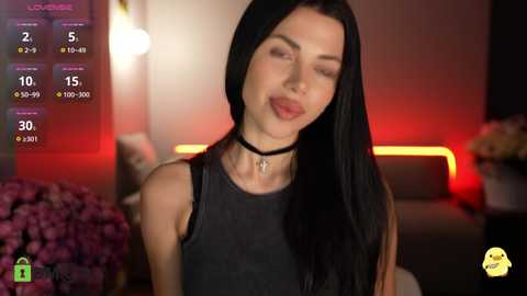 Video of a slim, fair-skinned woman with long black hair, wearing a black sleeveless top and choker, smiling in a dimly lit room with pink flowers and glowing red lights in the background.