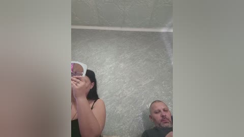 Video of a woman taking a selfie in a bathroom with textured gray walls, reflecting in the mirror. She has long black hair, is wearing a black top, and is holding a phone.