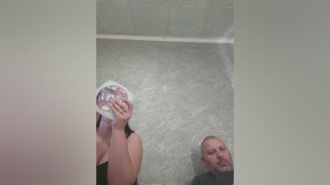 Video of a woman with medium skin tone, wearing a black top, holding a white plastic bag, standing next to a bald man in a dark shirt, both looking down at the carpeted floor.