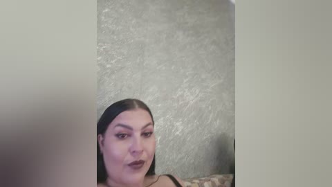 Video of a young woman with long, straight black hair, wearing dark lipstick, looking directly at the camera. She has a pale complexion and is wearing a dark top. The background is a textured, light gray wall.