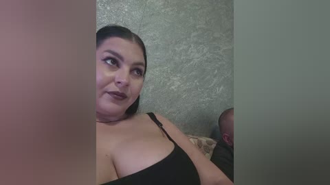 Video of a plus-sized woman with dark hair, wearing a black strapless top, sitting in a dimly-lit room with a textured grey wall behind her.