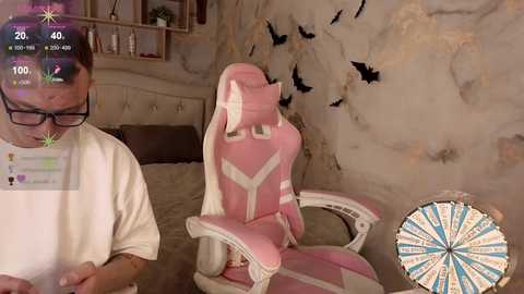 Video of a man in a beige t-shirt, sitting on a pink gaming chair with a digital overlay showing a dartboard, in a cozy, beige-themed room with bird decals.
