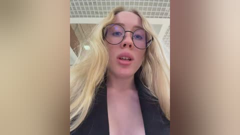 A video of a blonde woman with fair skin and blue eyes, wearing large round glasses and a black top, taken indoors with a tiled ceiling.