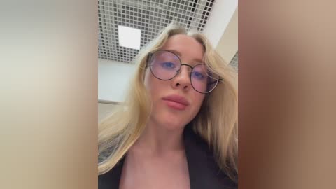 Video of a blonde woman with fair skin, wearing large, round glasses and a black top, taken from a low angle, showing her face and upper body. The background features a grid-patterned ceiling and white walls.