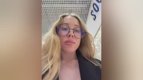 Video of a young Caucasian woman with blonde hair, wearing large, round glasses and a black top, standing in a modern, tiled bathroom.