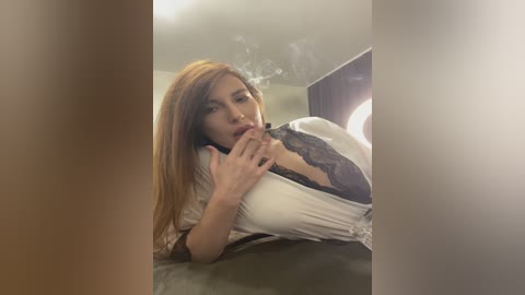 Video of a light-skinned woman with long, straight, light brown hair, wearing a sheer, white, lace-trimmed nightgown, lying on a bed with a dimly lit, blurred background, smoking a cigarette.