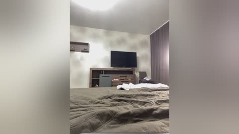 A video of a simple, modern bedroom with a queen-sized bed, beige quilt, and dark wooden dresser holding a TV and air conditioner. White walls, light brown curtains, and a single window.