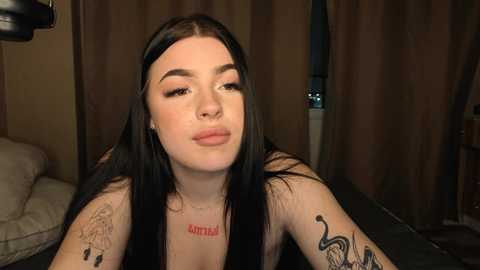 Video of a young woman with long black hair, fair skin, and multiple tattoos, including a snake on her arm. She wears a black top and sits in a dimly lit room with brown curtains and a lamp.