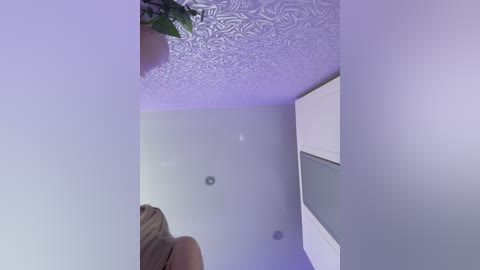 A video showing a woman's blonde hair from behind, standing in a room with light purple walls and a textured ceiling. The room has two small ceiling lights and a white door on the right.