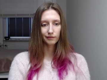 Video of a young Caucasian woman with long, straight, light brown hair transitioning to vibrant pink at the tips. She wears a white, textured sweater, standing in a simple, softly lit room with white walls and a window with partially drawn blinds.