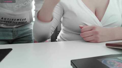 Video of a white woman in a white tank top and gray leggings, pulled down slightly to reveal her small breasts, sitting at a white desk.