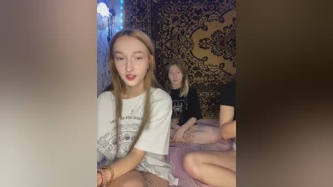 Video of two young women, one with long blonde hair in a white t-shirt, the other with straight brown hair in a black shirt, sitting on a bed with patterned curtains in the background.