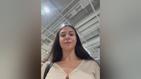Video of a young woman with long, dark hair, fair skin, and a neutral expression, standing in an industrial setting with a white ceiling and exposed pipes. She wears a cream-colored top.