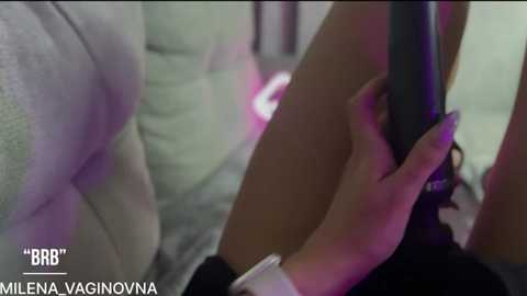 Video showing a close-up of a person's hand holding a black vibrator, with a blurry background featuring a couch and soft lighting. Text in the bottom left reads \"MILENA VAGINOVNA.\