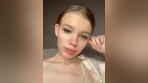 Video of a young Caucasian woman with light skin, straight brown hair, and wearing a white shirt, looking tired with her hand covering her eye.