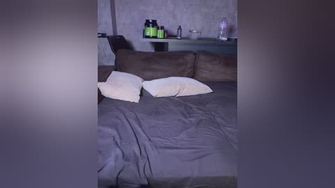 A video of a dimly lit bedroom with a bed covered in gray sheets, two beige pillows, and a brown headboard. A wooden dresser holds a bottle of water, a green bottle, and a black bottle, against a textured, light gray wall.