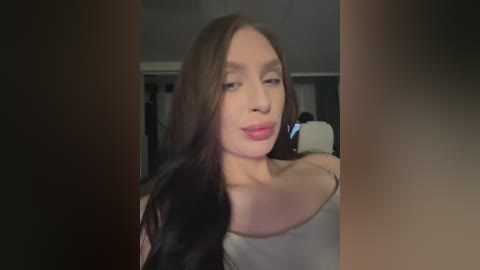 A video of a young woman with fair skin, long brown hair, and a white off-the-shoulder top. She has a serene expression, set against a dimly lit, indistinct background.