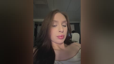 A video of a young woman with long brown hair, fair skin, and a neutral expression, wearing a white off-shoulder top, captured indoors with a dimly lit, modern room in the background.