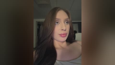 Video of a young woman with long brown hair, light skin, and pink lipstick, wearing a white off-shoulder top, taken indoors with blurred background.
