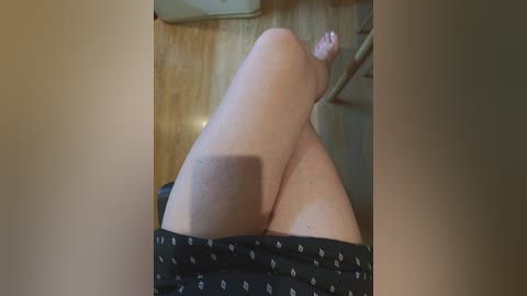 Video of a light-skinned person's legs, wearing black shorts, reclining on a wooden floor. The background shows a partially visible staircase and a beige wall.