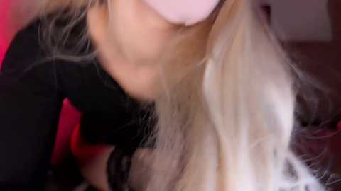 Close-up video of a woman with long, wavy blonde hair, wearing a black top, leaning forward. The background is blurred, highlighting her face and hair.