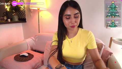 A video of a Latina woman with long black hair, wearing a yellow crop top, sitting on a pink couch in a cozy, dimly lit living room.